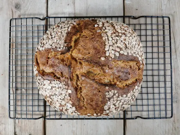 Ein rustikales Porridge Brot mit Haferflockenkruste, das auf einem schwarzen Gitter auf Holzdielen abkühlt.